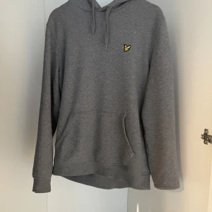 Grå hoodie från Lyle & Scott - Säljer en stilren grå hoodie från Lyle & Scott med klassisk gul logga på bröstet. Tröjan har huva med dragsko och en stor magficka. Perfekt för en avslappnad look.