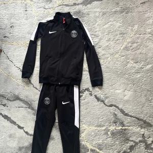 Svart träningsjacka från Nike PSG - Svart träningsjacka från Nike med PSG-logga på bröstet och vit panel längs axlarna. Jackan har dragkedja framtill, ståkrage och två fickor. Perfekt för sportiga tillfällen och har en modern, sportig look.