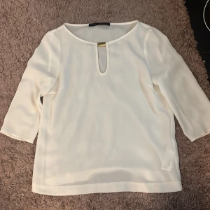 Vit blus med gulddetalj - Säljer en stilren vit blus med trekvartsärm och snygg gulddetalj vid halsringningen. Blusen har en liten öppning framtill som ger en elegant touch. Perfekt att matcha med både jeans och kjol.
