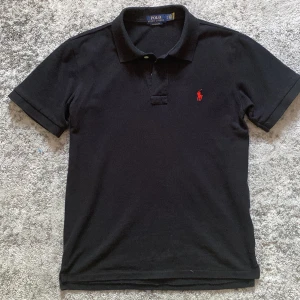 Svart pikétröja från Polo Ralph Lauren - Svart pikétröja från Polo Ralph Lauren med röd logga på bröstet. Custom slim fit, använd enstaka gång. Hör av er vid funderingar!