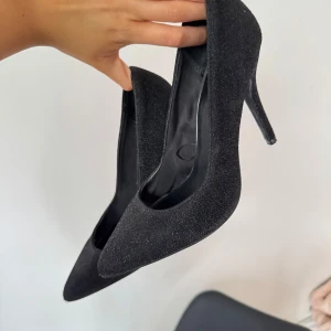 Svarta glittriga pumps från Bershka - Säljer ett par svarta pumps från Bershka med glittrig yta. Skorna har spetsig tå och hög klack, perfekta för att lyfta din outfit. Klassisk och elegant design med platt sula framtill.