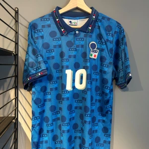 Italien retro fotbollströja nummer 10 - Blå kortärmad fotbollströja med mönster och nummer 10 på bröstet. Krage med knappar och detaljer i rött, vitt och grönt. Tryck 'R. Baggio' på ryggen. Perfekt för dig som gillar klassiska landslagströjor. Helt ny med tagg en replika