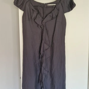 Mörkgrå klänning med volang - Säljer en elegant mörkgrå knälång klänning från Part Two. Klänningen har korta ärmar, volangdetalj framtill och stängs med dragkedja i ryggen. Perfekt för dig som gillar stilrena och feminina plagg.