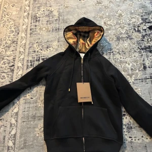 Svart hoodie från Burberry med dragkedja - Säljer en svart hoodie från Burberry med dragkedja och klassiskt rutigt foder i huvan. Tröjan har fickor framtill och justerbar huva med snören. Perfekt för dig som gillar stilrena märkesplagg.