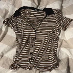 Brun och vit randig skjorta från BikBok - Kortärmad skjorta från BikBok i brun, vit och svart randigt mönster. Skjortan har svarta knappar framtill och en svart krage som kontrast. Materialet är ribbat och stretchigt, vilket ger en bekväm passform. Det är storlek S, har använt denna ungefär 2-3 gånger, så den är som ny 