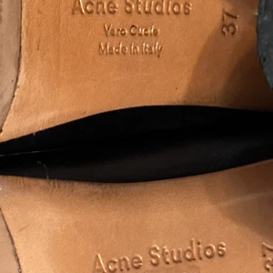 Svarta mules med röd klack från Acne Studios - Säljer ett par svarta mules från Acne Studios i äkta skinn med rund tå och bred röd klack. Skorna är tillverkade i Italien och kommer med original dustbags. Perfekta för dig som vill ha något unikt och stilrent.