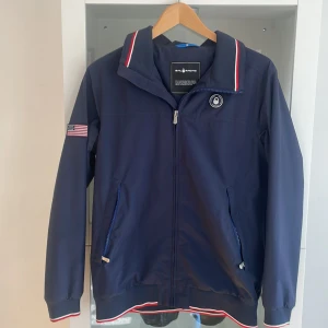 Marinblå vindjacka med USA-detaljer från Sail Racing - Snygg marinblå vindjacka från Sail Racing med dragkedja, USA-flagga på ärmen och trycket 'USA 77' på ryggen. Fodret i luvan har amerikanskt flaggmönster och jackan har röda och vita detaljer vid krage och muddar.