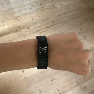 Svart monogram armband från Louis Vuitton - Stilrent svart armband från Louis Vuitton med ikoniska LV-loggan i silver på framsidan. Perfekt accessoar för att lyfta din outfit med en lyxig touch. Köpt på louis Vuitton butiken i Rom för 3500 Kr så har kvitto och allt og,  Inprincip helt nytt.