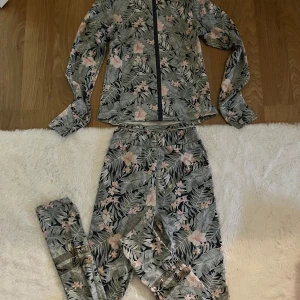 Blommigt tränings set med matchande tröja - Säljer ett tränings set  med grå bas och blommigt mönster i rosa och grönt. Byxorna har resår i midjan och är i ett mjukt material, perfekta för en avslappnad stil. Matchande tröja med dragkedja ingår.