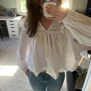 Vit blus med volang och spetsdetaljer - Säljer en luftig vit blus med v-ringning, volangdetaljer och spetsinslag vid halsen. Blusen har långa, vida ärmar med resår vid handlederna och en lös passform. Perfekt för vår och sommar!