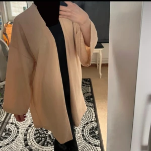 Beige overshirt med vida ärmar - Säljer en beige overshirt med lös passform och trekvartsärmar. Jackan är öppen framtill och har en stilren, minimalistisk design. Perfekt att slänga över en t-shirt eller tröja för en avslappnad look.
