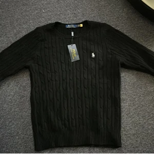 Svart kabelstickad tröja från Polo Ralph Lauren - Säljer en klassisk svart kabelstickad tröja från Polo Ralph Lauren med rund halsringning och broderad logga på bröstet. Tröjan har långa ärmar och ribbade muddar. Perfekt för en stilren look.