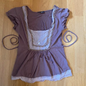 Ljusbrun blus med spetsdetaljer - Supersöt ljusbrun blus med spetsdetaljer vid halsringning och nederkant. Blusen har korta ärmar med rynk, knytband i midjan och dekorativ knapp framtill. Perfekt för dig som gillar romantisk stil.