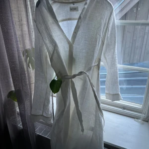Vit linnéklänning från Lin Living - Säljer en vit långärmad klänning i linne från Lin Living. Klänningen har knappar framtill, ett knytband i midjan och en avslappnad passform. Perfekt för varma dagar eller som strandplagg. 100% linne och är helt oanvänd! Nypris: 1850kr
