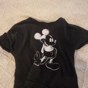 Svart Mickey Mouse t-shirt från Disney - Svart t-shirt från Disney med stor vit tryck av Mickey Mouse på framsidan. Klassisk passform och korta ärmar, perfekt för dig som gillar retrostil och tecknade motiv.