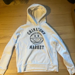 Vit hoodie från Chinatown Market - Vit hoodie med svart Chinatown Market-tryck och smiley på bröstet. Klassisk huva med dragsko och känguruficka framtill. Perfekt för en avslappnad streetwear-look.