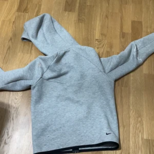 Grå hoodie från Nike med svarta detaljer(Nike tech) - Säljer en grå hoodie från Nike med svarta detaljer och dragkedja framtill. Tröjan har huva, två fickor och en extra ficka på ena ärmen. Perfekt för en sportig och avslappnad stil. Den har bara lite skråmor på armbågarna annars är den helt okej