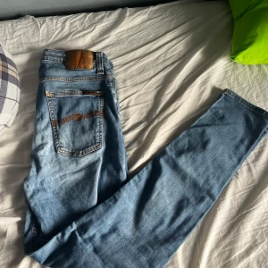 Ljusblå jeans från Nudie Jeans - Säljer ett par ljusblå jeans från Nudie Jeans med klassiska orange sömmar och broderad bakficka. Jeansen har en rak passform och är tillverkade i mjukt denimtyg. Perfekta för en avslappnad stil.