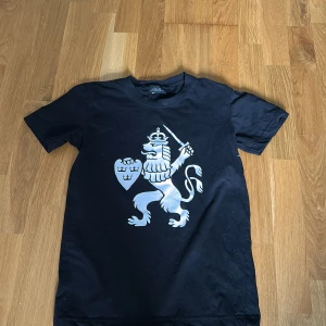 Svart ifk tröja  - Svart t-shirt med stort vitt lejontryck på framsidan. Klassisk rund hals och korta ärmar. Perfekt för dig som vill visa ditt stöd för Sverige på ett stilrent sätt.