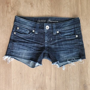Mörkblå jeansshorts från Guess Premium - Snygga mörkblå jeansshorts från Guess Premium med fransiga benslut och klassisk femficksdesign. Shortsen har låg midja och är tillverkade i stretchigt material med 60% bomull och 40% polyester. Perfekta för sommardagar!
