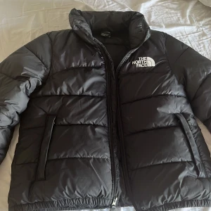 Svart dunjacka från The North Face - Säljer en svart dunjacka från The North Face med hög krage och dragkedja. Jackan har två sidofickor med dragkedja och logga på bröstet och ryggen. Perfekt för kyliga dagar och riktigt skön att bära. ( kan också prata om och sänka priset )