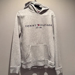 Grå hoodie från Tommy Hilfiger - Grå hoodie med broderad Tommy Hilfiger-logga och flagga på bröstet. Klassisk känguruficka framtill och huva. Mjuk och bekväm modell, perfekt för vardagsbruk. Storlek M men passar även S. Priset går att diskutera.