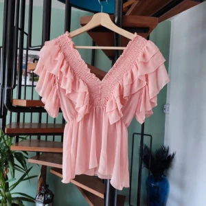 Rosa volangblus med v-ringning - Söt rosa blus med djup v-ringning och flera lager volanger längs axlar och byst. Blusen har korta ärmar och smockad överdel som ger en feminin och luftig känsla. Perfekt för vår och sommar.