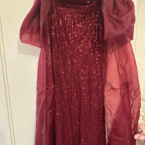 Vinröd paljettklänning med puffärmar - Säljer en vinröd långklänning med glittriga paljetter och puffiga kortärmar. Klänningen har en elegant och festlig look med transparent tyg över axlarna och ärmarna. Perfekt för bal eller festliga tillfällen.