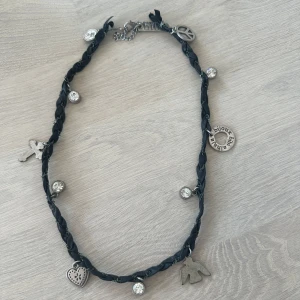 Svart flätat halsband med berlocker - Unikt svart flätat halsband med flera silverfärgade berlocker, bland annat hjärta, fågel, peace-märke och glittriga stenar. Perfekt accessoar för att ge din outfit en personlig touch.