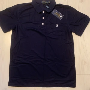 Mörkblå sport pikétröja från Polo Ralph Lauren - Säljer en klassisk mörkblå sport pikétröja från Polo Ralph Lauren med vit broderad logga på bröstet. Tröjan har korta ärmar, krage och tre knappar framtill. Perfekt för en stilren look.