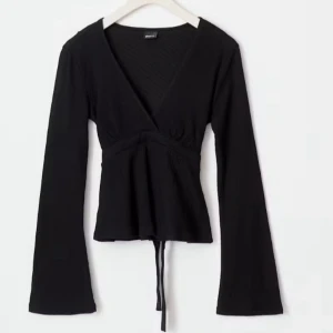 Svart omlottopp med knytband - Säljer en svart omlottopp med långa ärmar och v-ringning. Toppen har knytband i midjan och är tillverkad i ett mjukt, stretchigt material. Perfekt att styla med jeans eller kjol för en trendig look. Endast använd en gång. Skriv för mer bilder.