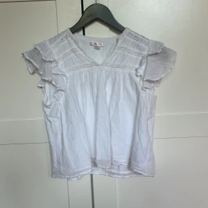 Vit blus med volang från Gina Tricot - Säljer en söt vit blus från Gina Tricot med v-ringning och volangdetaljer på axlarna. Perfekt för vår och sommar! Säljer pga att den fort blev för liten, använd kanske max 4 ggr så den är som ny!