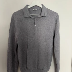 Grå pikétröja i merinoull från John Henric - Säljer en stilren grå långärmad pikétröja från John Henric. Tröjan är tillverkad i mjuk merinoull och har klassisk krage samt knäppning framtill. Perfekt för en clean och avslappnad look.