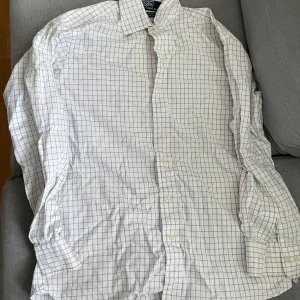 Vit rutig skjorta från Polo Ralph Lauren - Vit skjorta med tunna blå rutor från Polo Ralph Lauren, modell Curham Classic Fit. Skjortan har lång ärm, klassisk krage och knäppning framtill. Perfekt för en clean och stilren look.