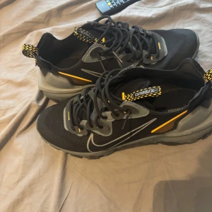 Svarta Nike sneakers med grå och gula detaljer - Snygga svarta Nike sneakers med grå och gula detaljer. Skorna har snörning, rund tå och platt sula. Perfekta för dig som vill ha en sportig och modern look. Nike-loggan syns tydligt på sidan och hälen har ett coolt mönster.