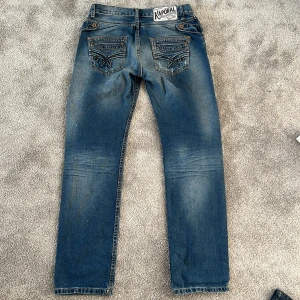 Blå jeans från Kaporal - Säljer ett par klassiska blå jeans från Kaporal med snygga slitningar och broderade bakfickor. Jeansen har raka ben och normal passform, samt detaljerade sömmar och en tydlig Kaporal-lapp bak i midjan.