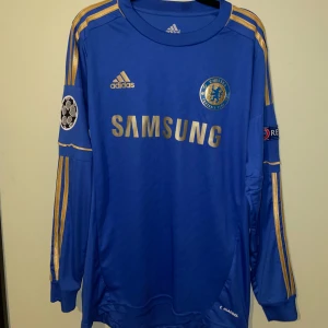 Chelsea Hazard 17 långärmad 12/13 - Chelsea FC långärmad fotbollströja från Adidas med Hazard 17 på ryggen. Blå med guldiga detaljer, klubbmärke och Champions League-patch på ärmen. Tillverkad i lätt och ventilerande material, perfekt för dig som älskar fotboll och vill sticka ut på planen.