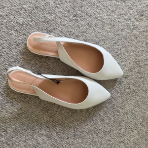 Vita ballerinaskor från Anna Field - Snygga vita ballerinaskor från Anna Field med spetsig tå och öppen häl. Skorna har en stilren design med slät ovandel och rem runt hälen. Perfekta för dig som gillar minimalistisk och fräsch stil.