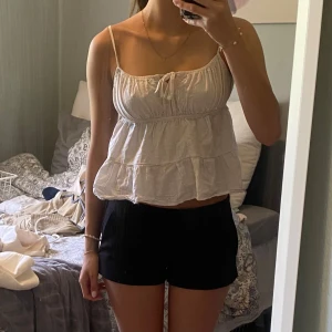 Ljusvit volangig linnetopp med knyt - Supersöt ljusvit linnetopp med tunna axelband och volanger. Toppen har en croppad passform och en liten knytning framtill. 