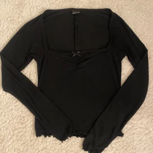 Svart långärmad topp med rosett - Supersöt svart långärmad topp från Gina Tricot med liten rosett och spetsdetalj vid urringningen. Toppen har croppad passform och vågig kant nertill för en extra trendig look. Perfekt att styla med jeans eller kjol för en cool vibe.