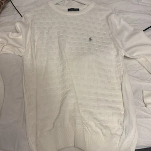 Vit stickad tröja Polo Ralph Lauren - Vit stickad tröja från Polo Ralph Lauren med diskret rutmönster och broderad logga på bröstet. Klassisk rund halsringning och ribbade muddar. Perfekt för en clean och stilren look. Kom med bud