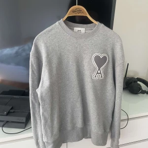 Grå sweatshirt från AMI Paris - Säljer en stilren grå sweatshirt från AMI Paris med deras ikoniska hjärta-A-logga broderad på bröstet. Tröjan har rund hals, ribbade muddar och är tillverkad i mjuk bomull. Perfekt för dig som gillar minimalistisk och trendig streetwear.