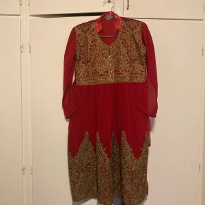 Röd långklänning med guld broderi - Unik röd långklänning med långa ärmar och v-ringning. Klänningen har detaljerat guld broderi över bröstet och längs nederkanten, samt knappar framtill. Tunt, lätt material som ger ett elegant fall. Perfekt för dig som vill sticka ut med en klassisk och lyxig look.