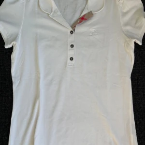 Vit pikéblus från Burberry Brit - Vit pikéblus från Burberry Brit med klassisk krage, tre knappar framtill och diskret broderad logga på bröstet. Kragen har en snygg detalj med Burberrys rutiga mönster på insidan. Står L men passar som M