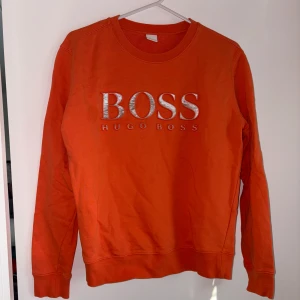 Orange sweatshirt från Hugo Boss - Snygg orange sweatshirt från Hugo Boss med stort silverbroderat BOSS-tryck på bröstet. Tröjan har rund halsringning, långa ärmar och är gjord i mjuk bomull. Perfekt för dig som vill sticka ut med en färgstark och trendig look.