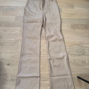 Beiga skinnbyxor från Madlady - Snygga högmidjade byxor i beige skinnimitation från Madlady. Byxorna har raka ben med lätt bootcut och klassisk femficksdesign. 