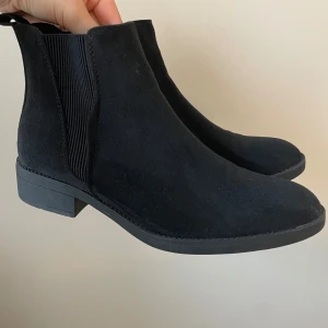 Svarta chelsea boots från About You - Stilrena svarta chelsea boots i mocka-look från About You. Skorna har elastiska paneler på sidorna, låg klack och rund tå. Perfekta för dig som vill ha en klassisk och enkel stil med edge. Snygga till både jeans och kjol. Stl 38. Sparsamt använda  
