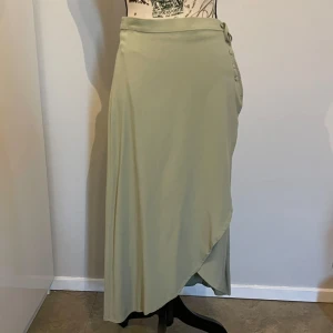 Ljusgrön omlottkjol med knyt - Supersnygg ljusgrön långkjol i omlottmodell med knyt och klädda knappar i sidan. Kjolen har en mjuk, flowy känsla och passar perfekt till sneakers eller sandaler. Enkel och stilren design som ger en chill men ändå trendig vibe.