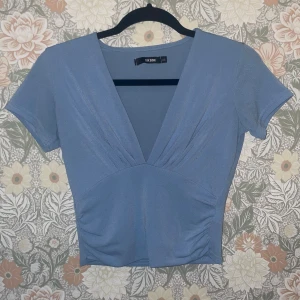 Blå v-ringad topp från Bik Bok - Snygg blå topp från Bik Bok med djup v-ringning och korta ärmar. Toppen har lätt rynkade detaljer framtill som ger en schysst form och sitter figurnära. Perfekt att styla med jeans eller kjol för en trendig look.