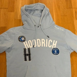 Ljusblå hoodie från Hoodrich - Snygg ljusblå hoodie från Hoodrich med broderade patches och vit logga på bröstet. Tröjan har huva med snörning och känguruficka framtill. 